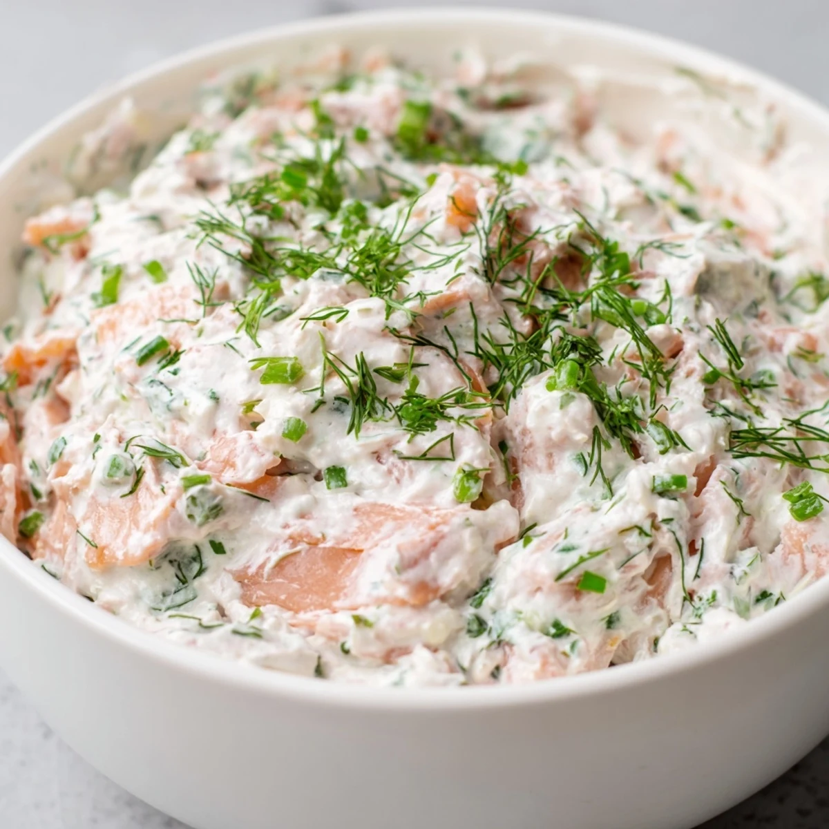 A bowl of creamy smoked salmon dip garnished with fresh dill and chives, served with crackers and cucumber slices.