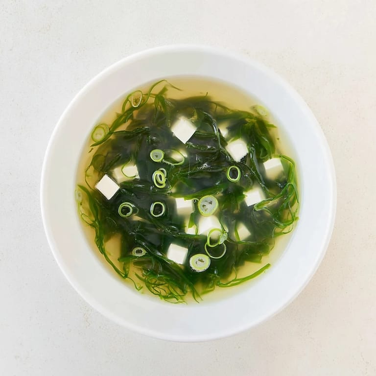 A spoon dips into savory wakame soup, revealing tender tofu and vibrant green seaweed in a fragrant, mineral-rich dashi broth.  